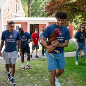 A group of people walking outside. There is a red barn in the background. One person is smiling, holding a rooster.