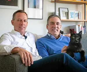 Two people are sitting inside on a couch, smiling, with a black cat on one person's lap.