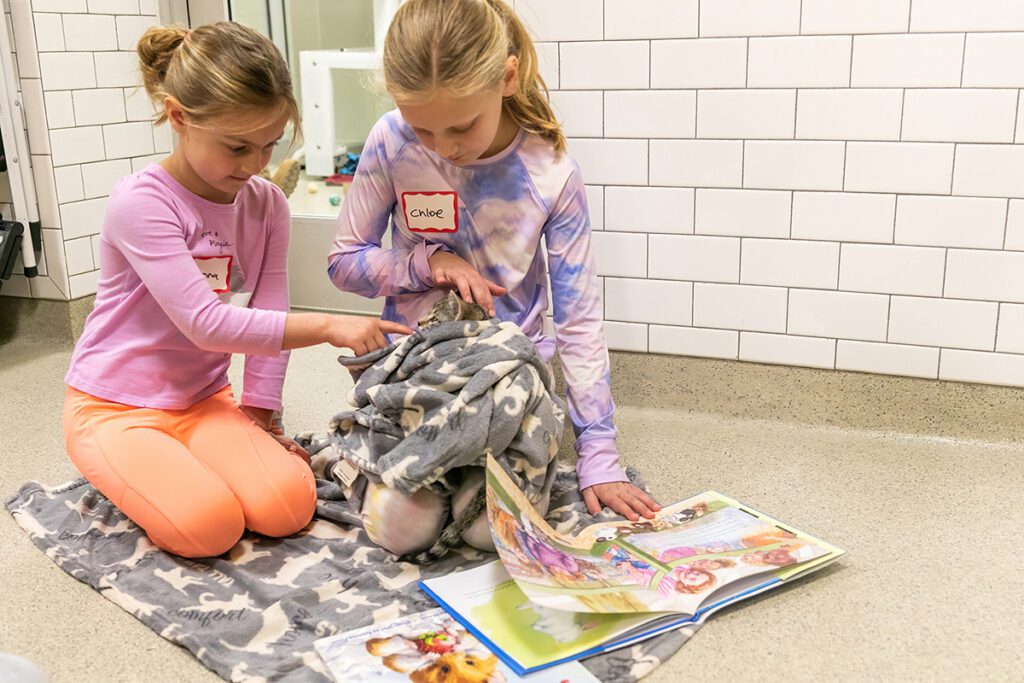 Two young girls sitting on the floor reading to a kitten who is laying in a blanket in one of the girl's laps. 