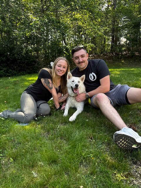 A large white dog lying down outside in grass, betwen two smiling people.