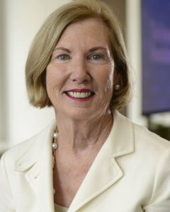 Portrait of a smiling woman with short blonde hair, red lipstick, pearl earrings, and a cream blazer.