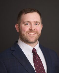 Professional headshot of a smiling bearded man in a navy suit, white shirt, and patterned burgundy tie against a dark background.