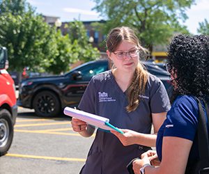 A smiling veterinary worker in scrubs holds a clipboard while speaking with a woman outdoors near parked cars.