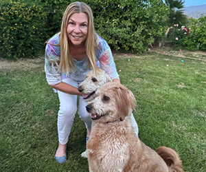Smiling woman kneels beside two golden dogs in a grassy garden.