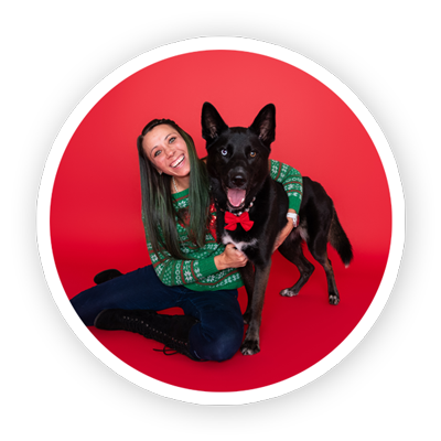 Smiling woman in a green holiday sweater kneels beside a black dog wearing a red bow tie, posed against a bright red backdrop in a festive portrait.