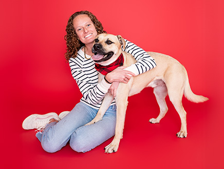 A smiling woman kneels beside and hugs a tan dog wearing a red bandana against a bright red backdrop, conveying warmth and affection.