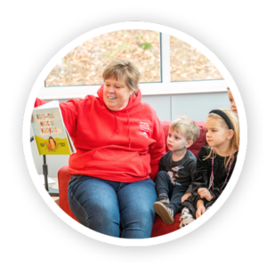 A smiling person in a red sweatshirt reads an illustrated book to young children seated beside her.